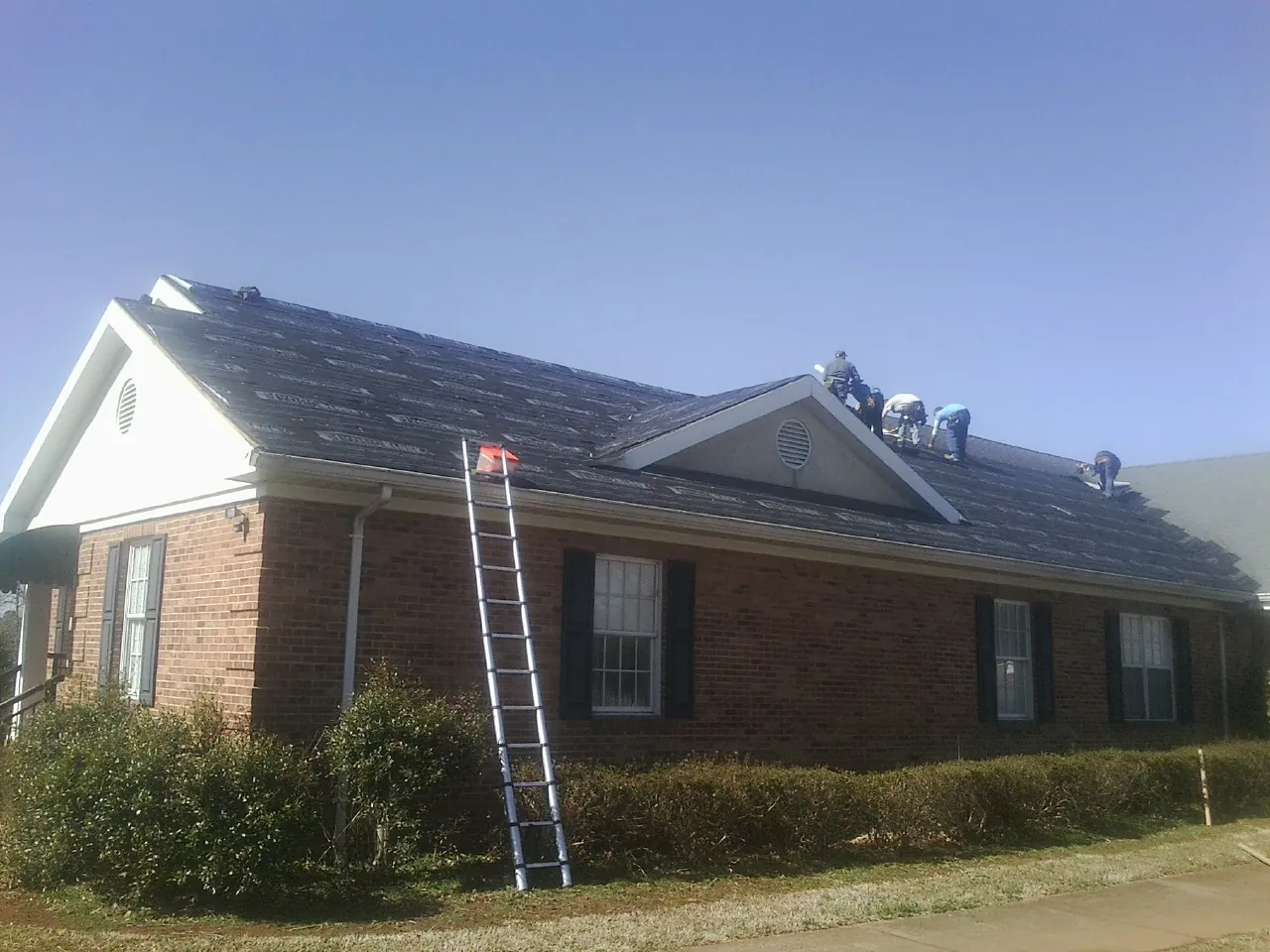 Crew tearing shingles off a brick ranch roof with a ladder set — Metal Roof Repair work by our crew in Stendal