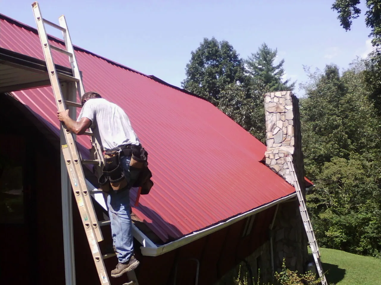 Roofer on a ladder beside a red metal roof and stone chimney — Soffit & Fascia Repair by Roofing Brothers in Stendal